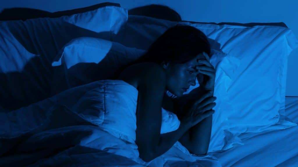 A woman is lying awake in bed, covering her forehead with her hands, looking stressed or suffering from insomnia.