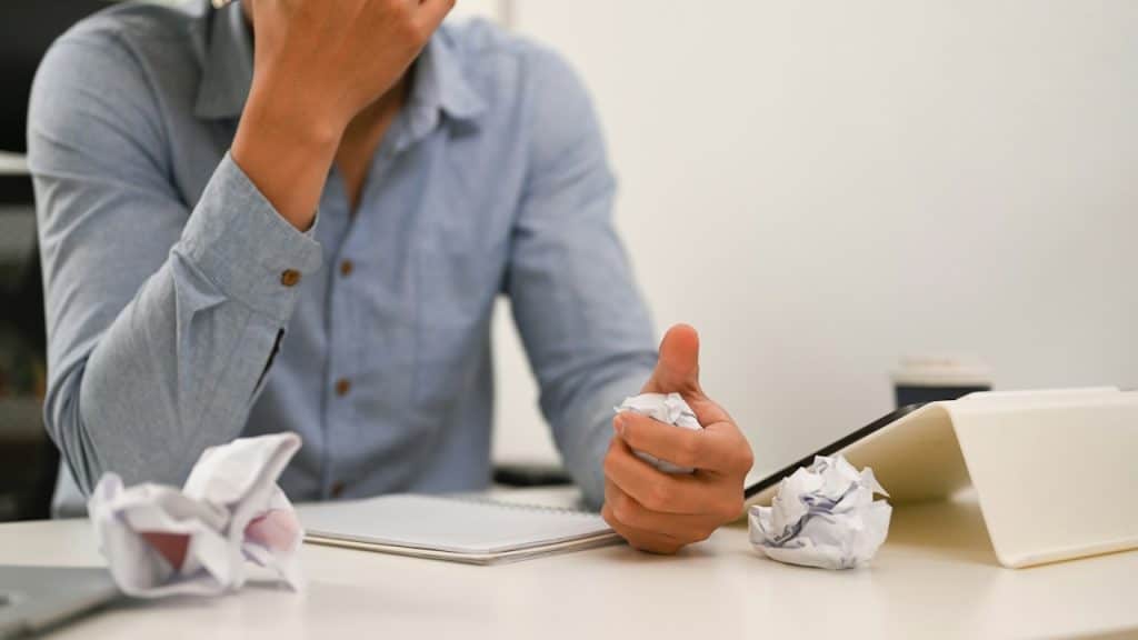 A man sitting at a white desk with his hand covering his face, looking stressed, while crumpling a piece of paper in his other hand.