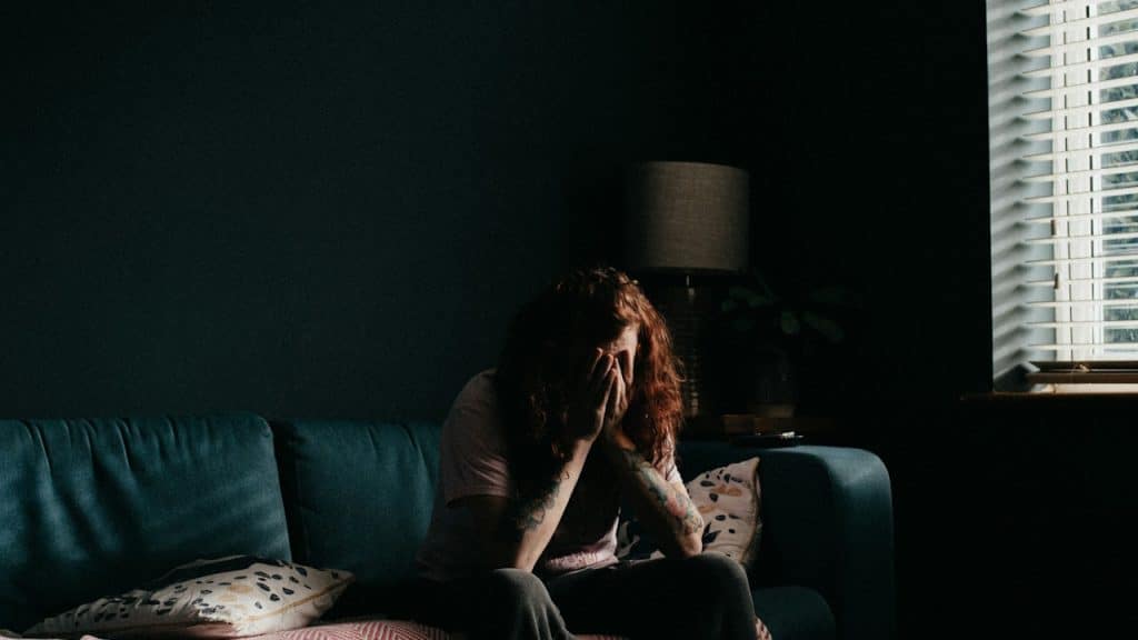 A distressed person with long red hair and tattoos sits on a teal sofa in a dimly lit room, covering their face with their hands.