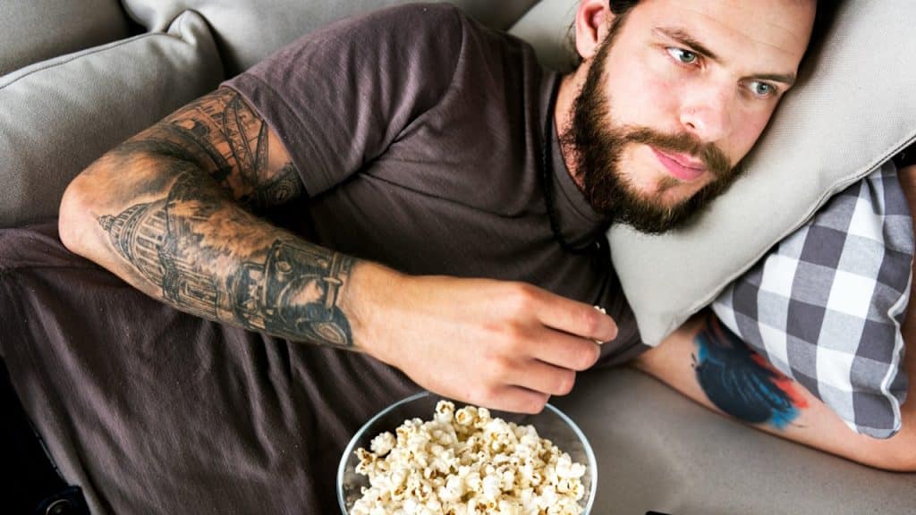 A man with a beard and tattoos lying on a couch with a bowl of popcorn.