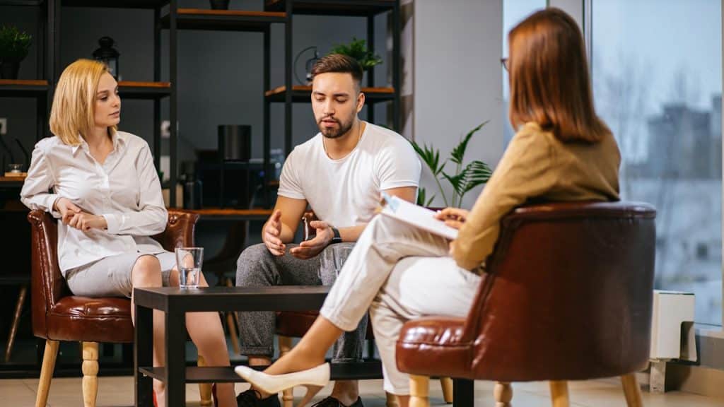 A couple sitting across from a female therapist, with the man speaking during a therapy session.