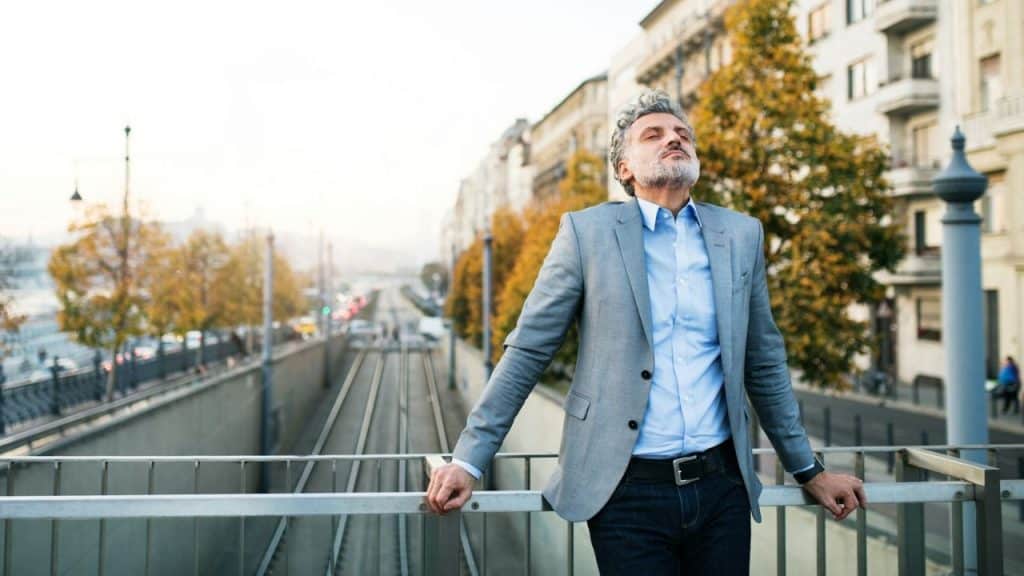 Grey-haired man in a grey blazer stands on a city bridge, eyes closed, looking up at the sky.