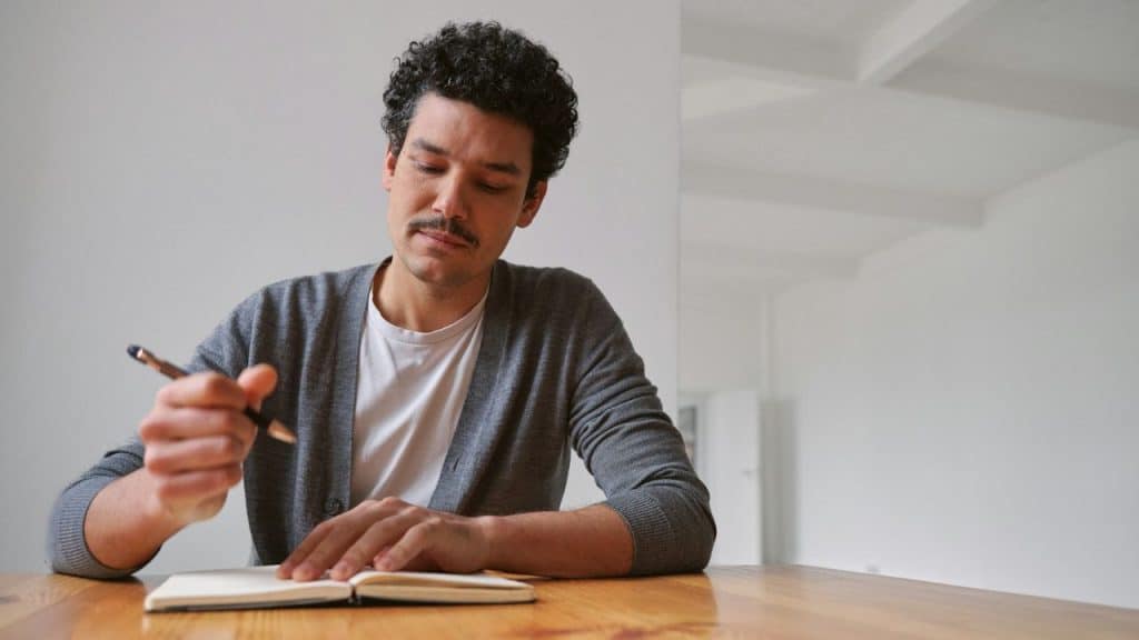 Man with a mustache and curly hair writing in a notebook at a wooden table.