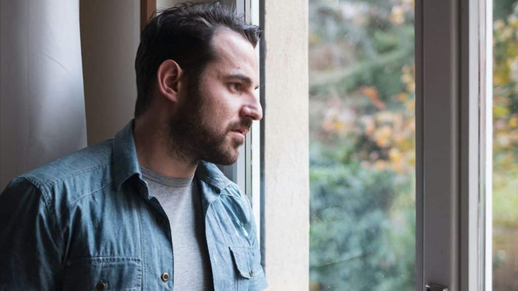 Serious man in a denim shirt looking out a window at blurred greenery and trees.