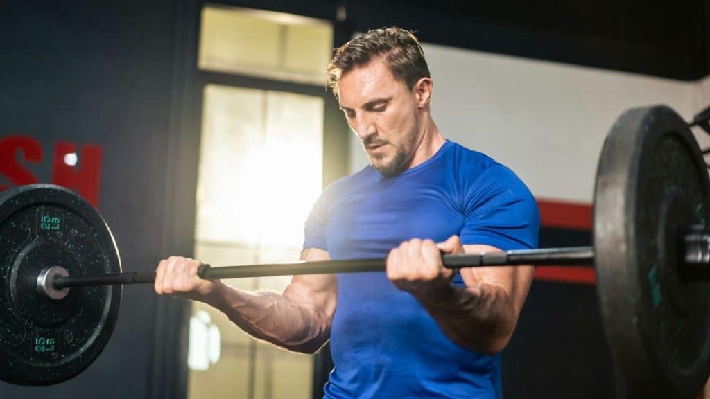 Muscular man in a blue shirt lifting a barbell in a gym setting.