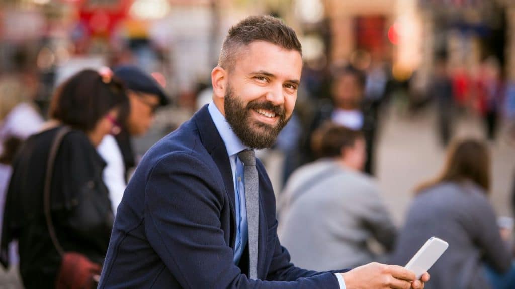 Smiling, bearded man in a suit and tie, holding a phone in a crowded outdoor area.