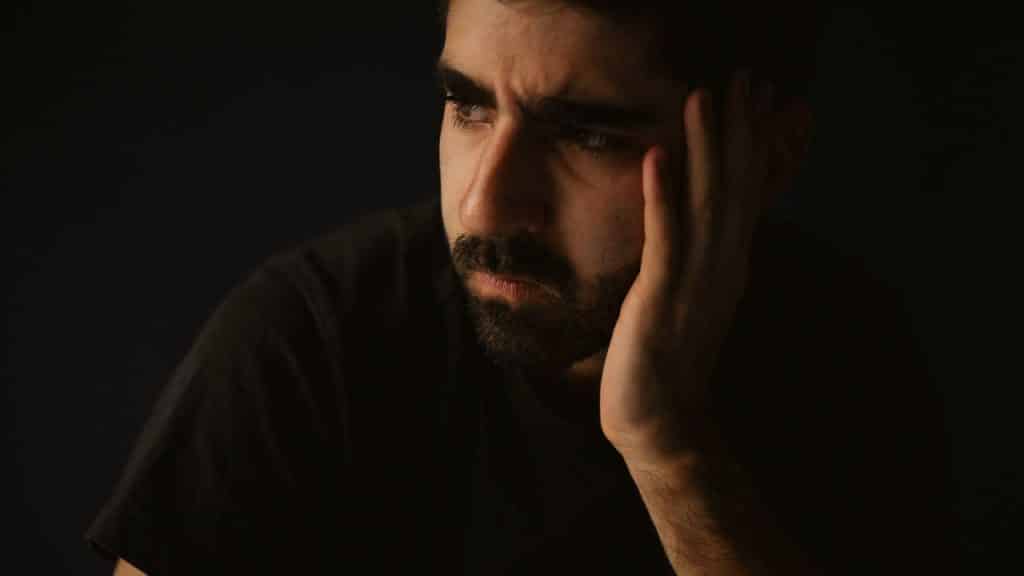 Solemn bearded man in shadow with his hand resting on his face, looking left.