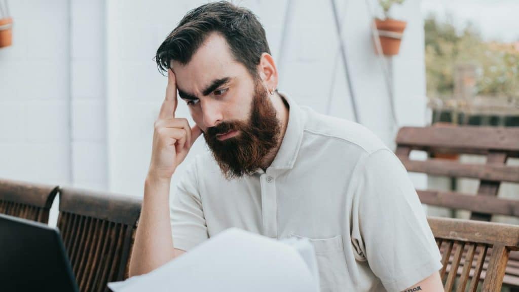 Bearded man looking stressed, holding his forehead while looking at papers and a laptop.