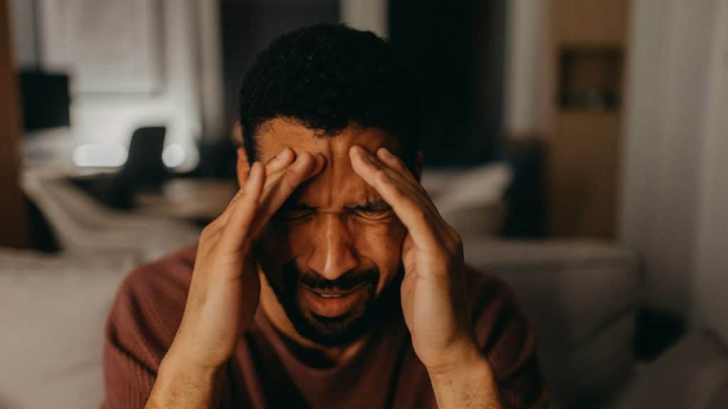 Distressed man with hands on his temples, eyes closed in a dimly lit indoor setting.
