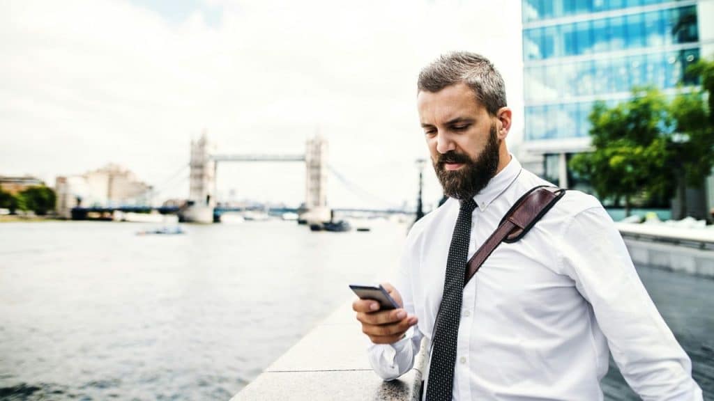 Bearded man in a white shirt and tie checking his phone near a river and bridge.
