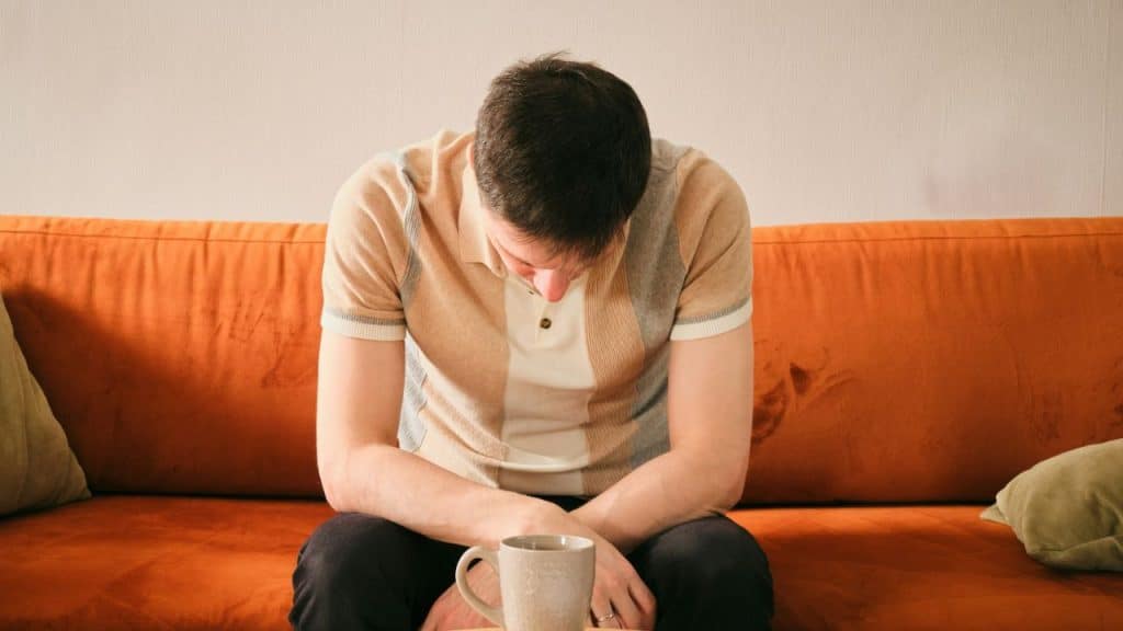 Man sitting on an orange couch with his head bowed, a mug is in front of him.