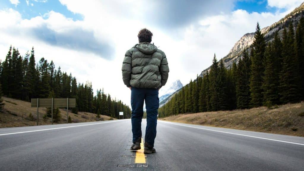 Man in a puffy jacket standing alone in the center of a road lined with trees and mountains.