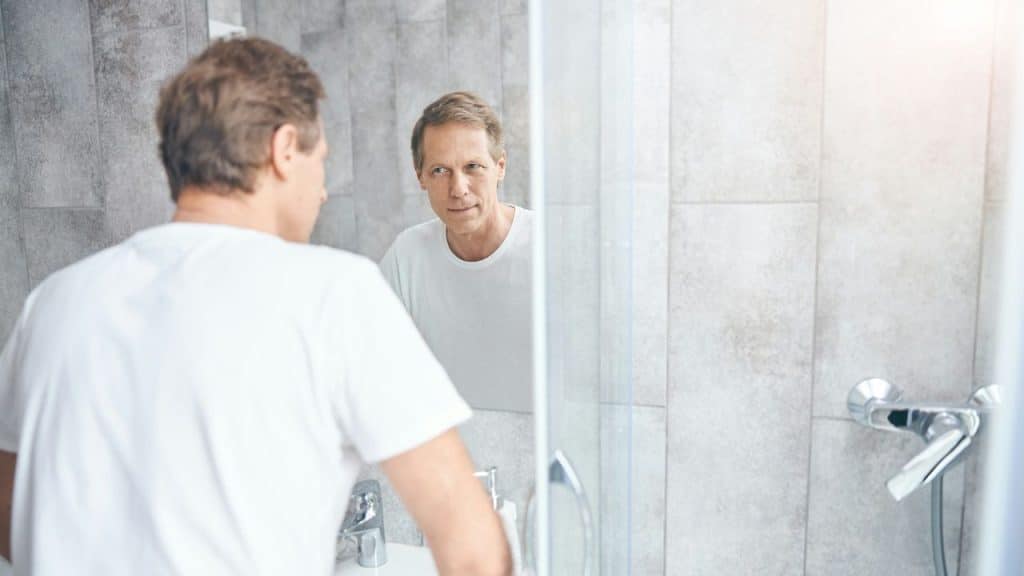 Man in a white shirt looking at his reflection in a bathroom mirror.