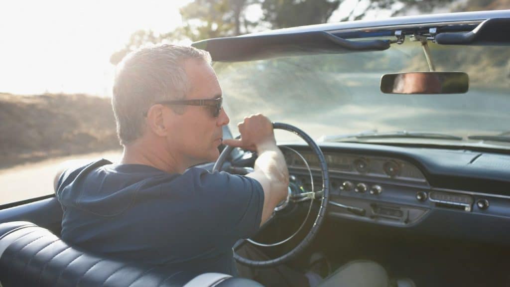 Middle-aged man wearing sunglasses, driving a convertible car on a sunny day.