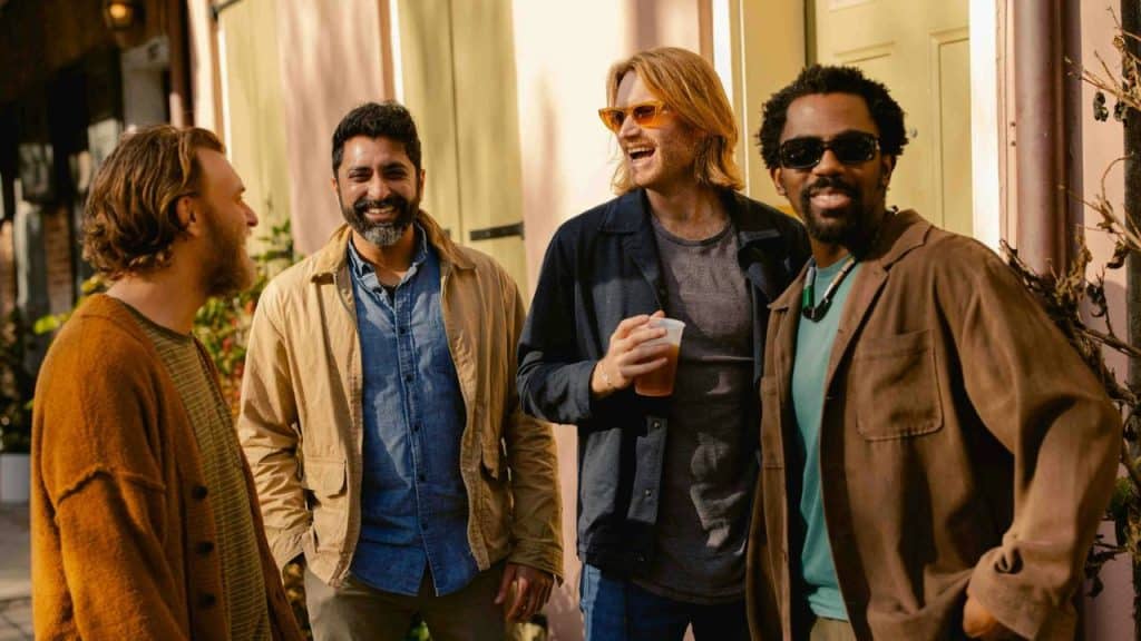 Four diverse men standing close together, smiling and chatting outdoors on a sunny day.