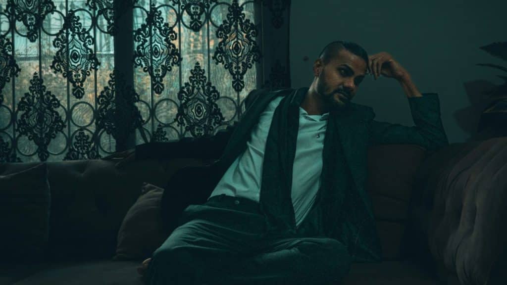 Solemn man in a suit sitting on a sofa in a dimly lit room with patterned curtains.