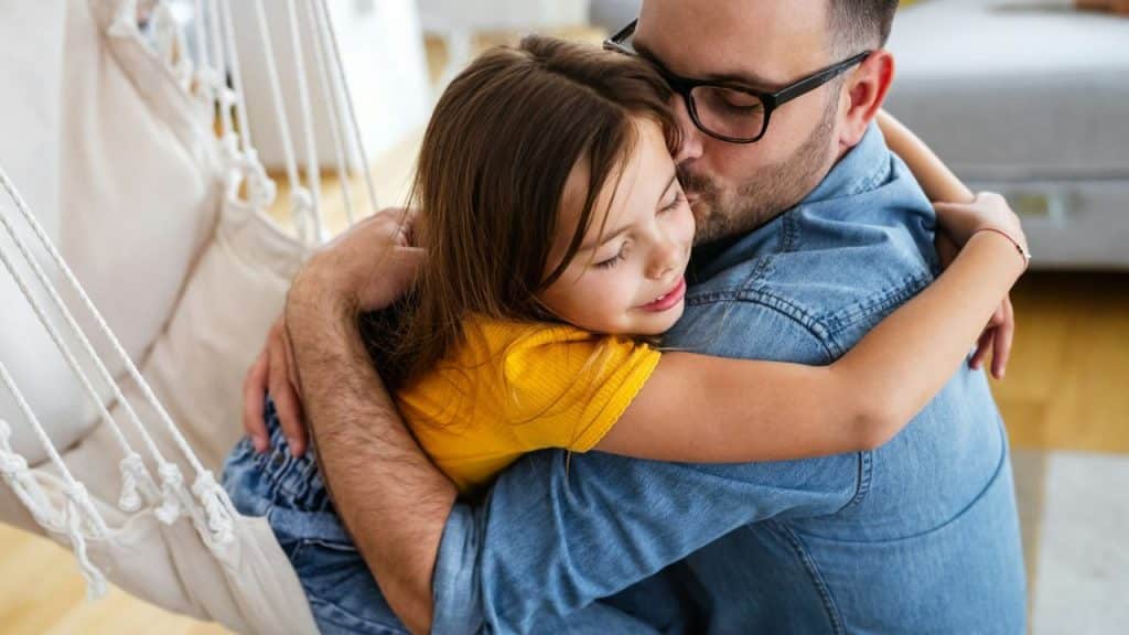 Father in glasses and a denim shirt hugging his young daughter in a bright indoor setting.