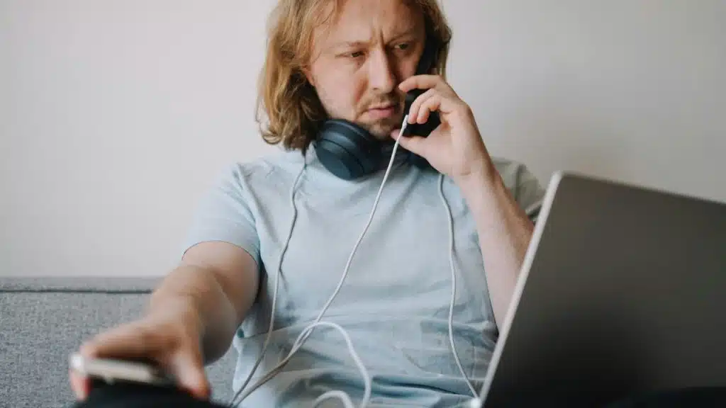 A long-haired man with headphones on his neck talks on a phone while also using a laptop.