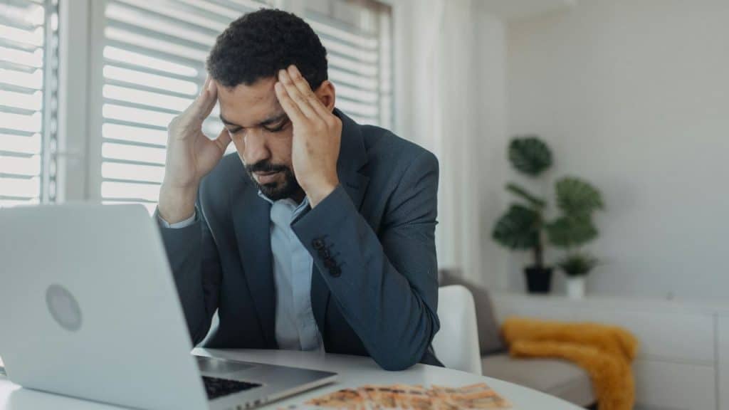 Stressed man in a suit holding his head while looking at a laptop and cash on a desk.