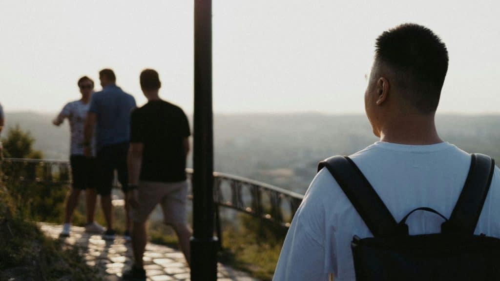 Man with a backpack stands looking at three blurred men on a sunlit outdoor path.