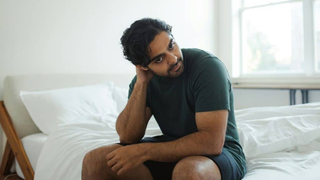 Man sitting on a bed with white sheets, hand behind his neck, looking up and right.