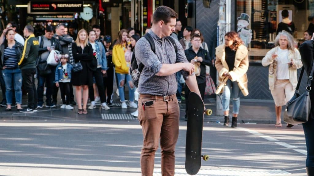 Man with a backpack and skateboard standing in a busy city street among a crowd.