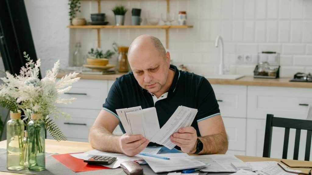 A serious, bald man sitting at a kitchen table reviewing several paper bills.