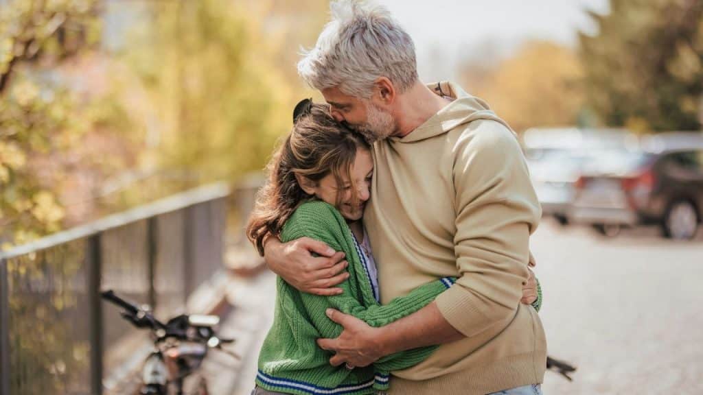 A loving father with gray hair hugging his daughter outdoors with a bicycle nearby.
