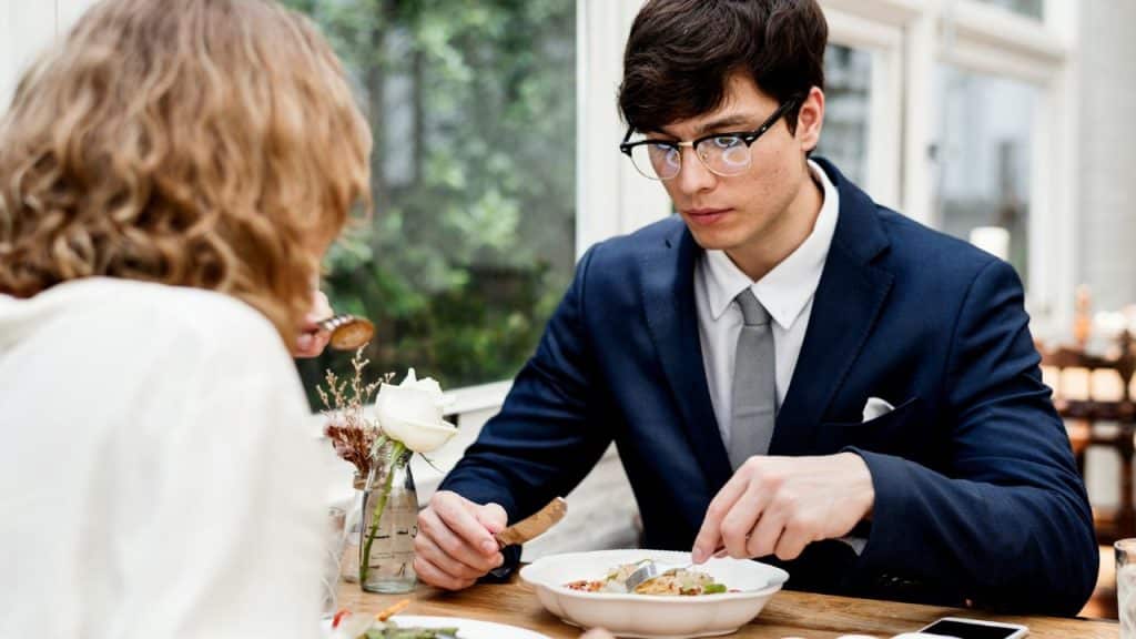 A serious young man in a suit and glasses eating a meal across a table from a woman.