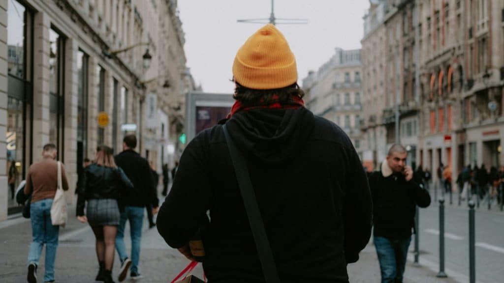 A man in an orange beanie and dark hoodie walks down a busy city street.