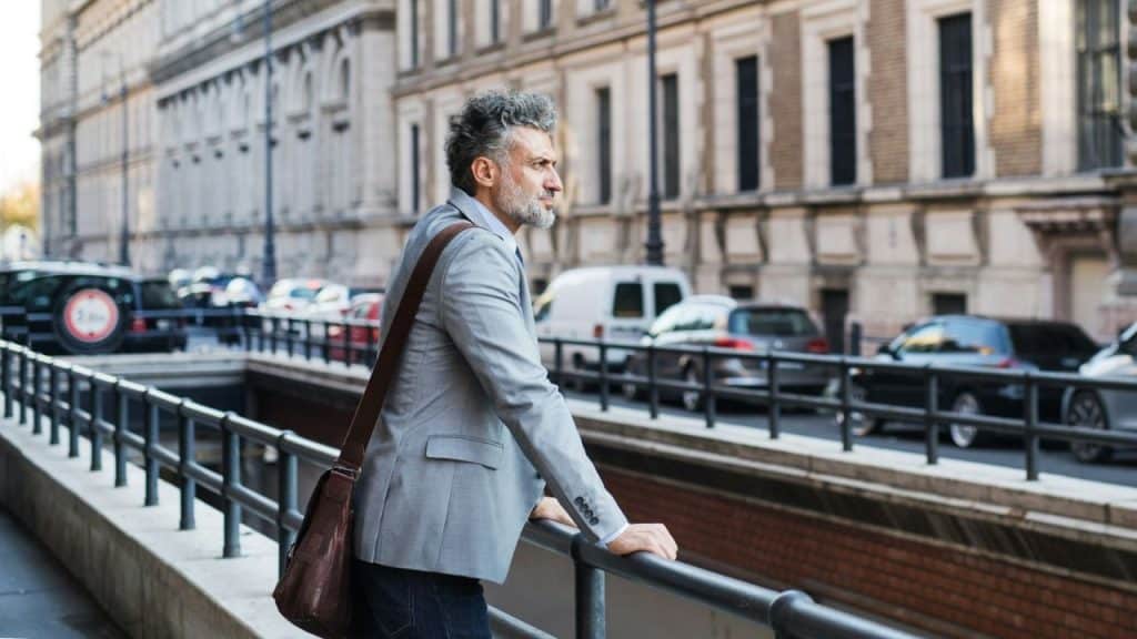 A middle-aged man in a gray jacket and shoulder bag looks pensively over a street railing.