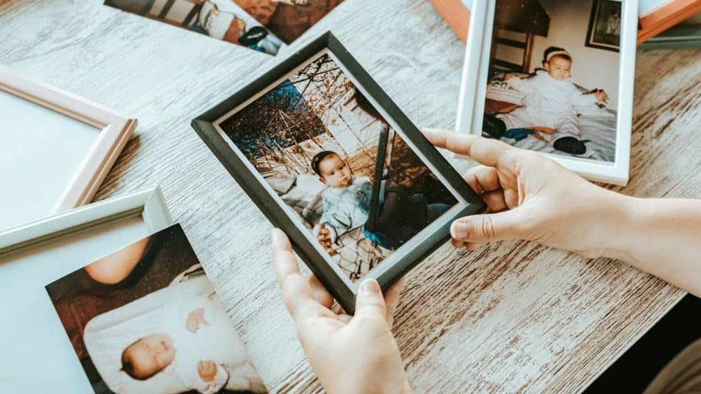Hands holding a framed photo of a baby on a wooden table with other photos.