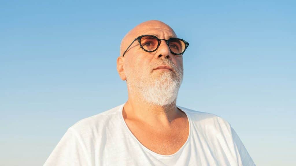 Close-up of a bald man with a white beard and dark-rimmed glasses looking up at the sky.
