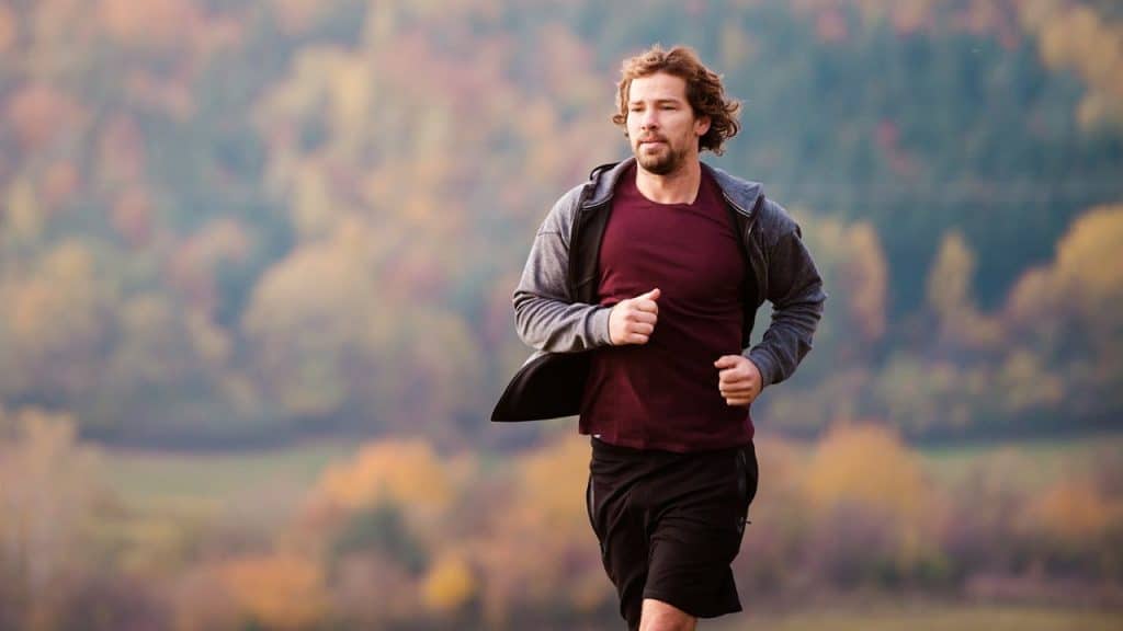 A bearded man in running clothes jogging outdoors with autumn foliage in the background.