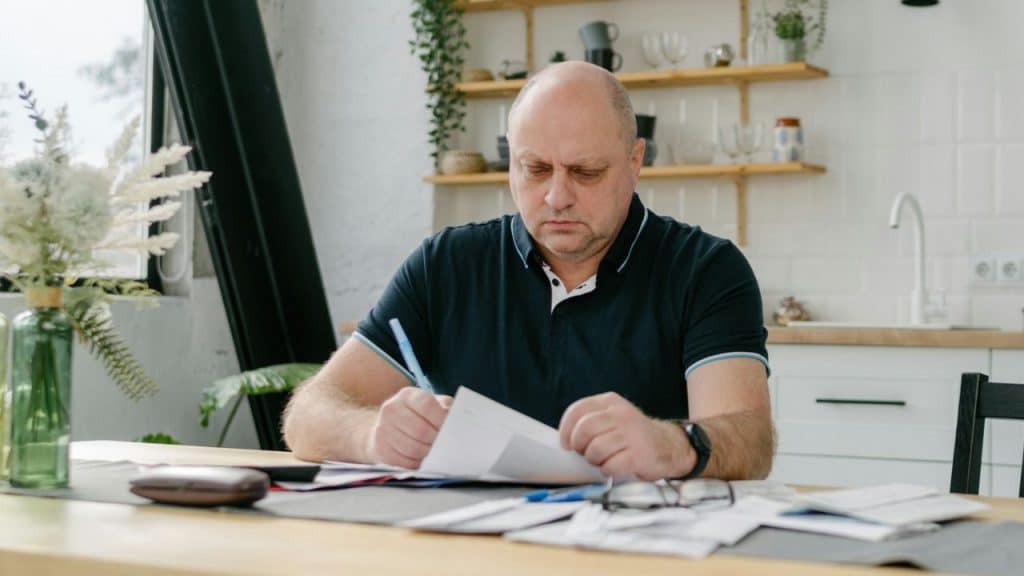 A bald, serious man sitting at a kitchen table reviewing documents and writing.