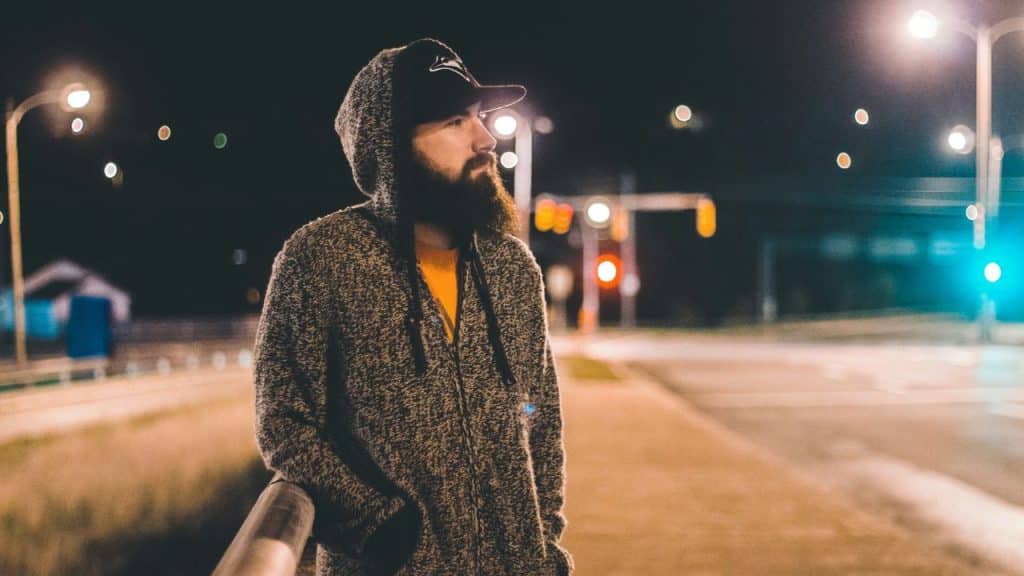 A bearded man in a hoodie and cap stands on a city street at night.