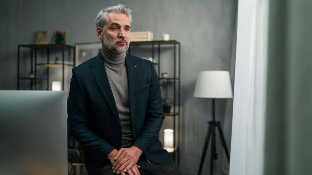 A handsome, gray-haired man in a turtleneck and suit jacket sitting in an office, looking pensive.