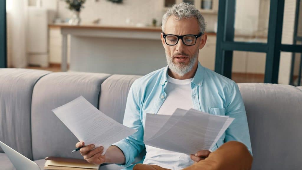 A gray-haired man in a blue shirt and glasses reading documents while sitting on a sofa.