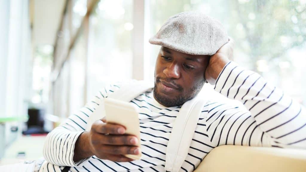 A man in a flat cap and a striped shirt looks at his smartphone indoors.