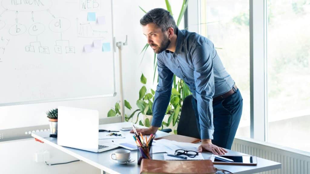 A serious man in a blue shirt leans over a desk looking at a laptop in an office.
