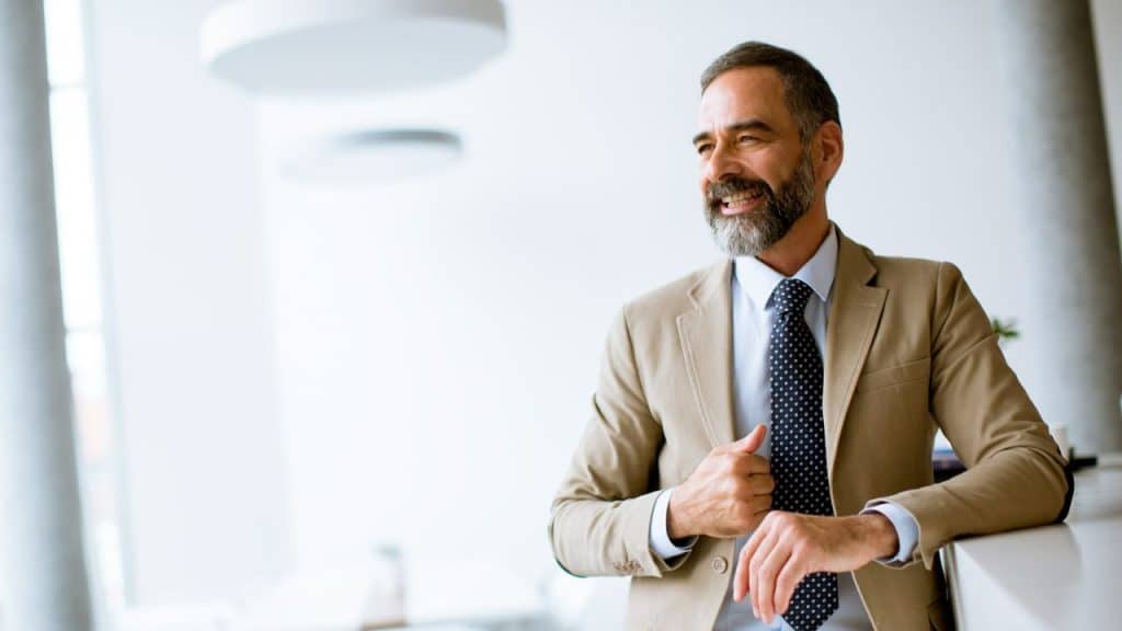 Smiling bearded man in a tan suit and tie stands leaning on a counter in a bright office.