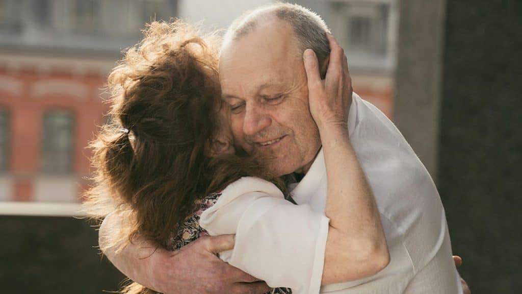 Close-up of an older couple warmly hugging outdoors, both smiling with eyes closed.