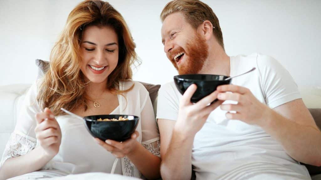 A happy couple laughing while eating breakfast cereal from black bowls in bed.