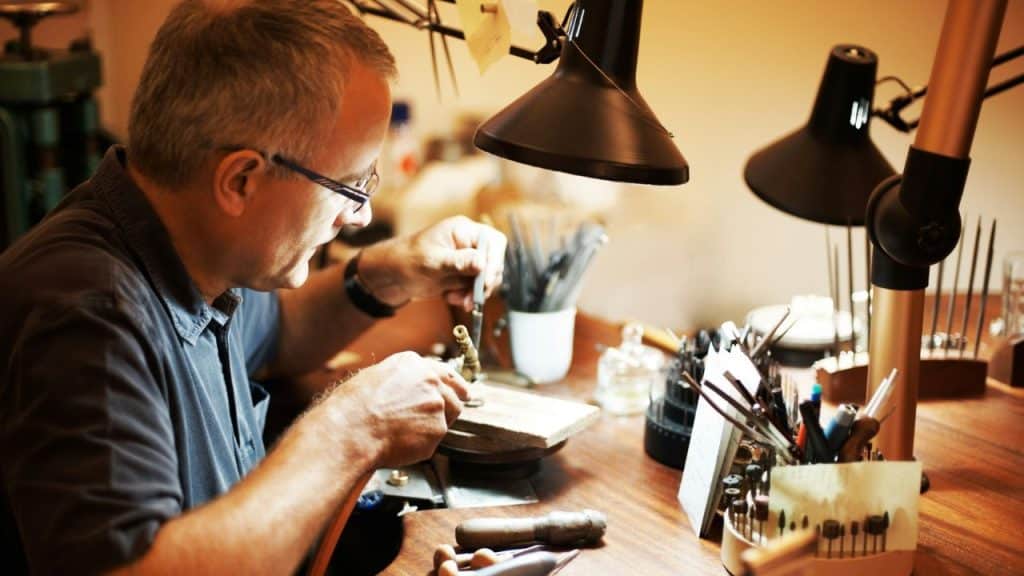 A man wearing glasses works intently on a small object at a workbench under bright lamps.