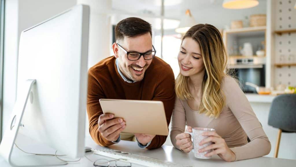 A smiling couple looking at a tablet while sitting at a computer desk in a kitchen.