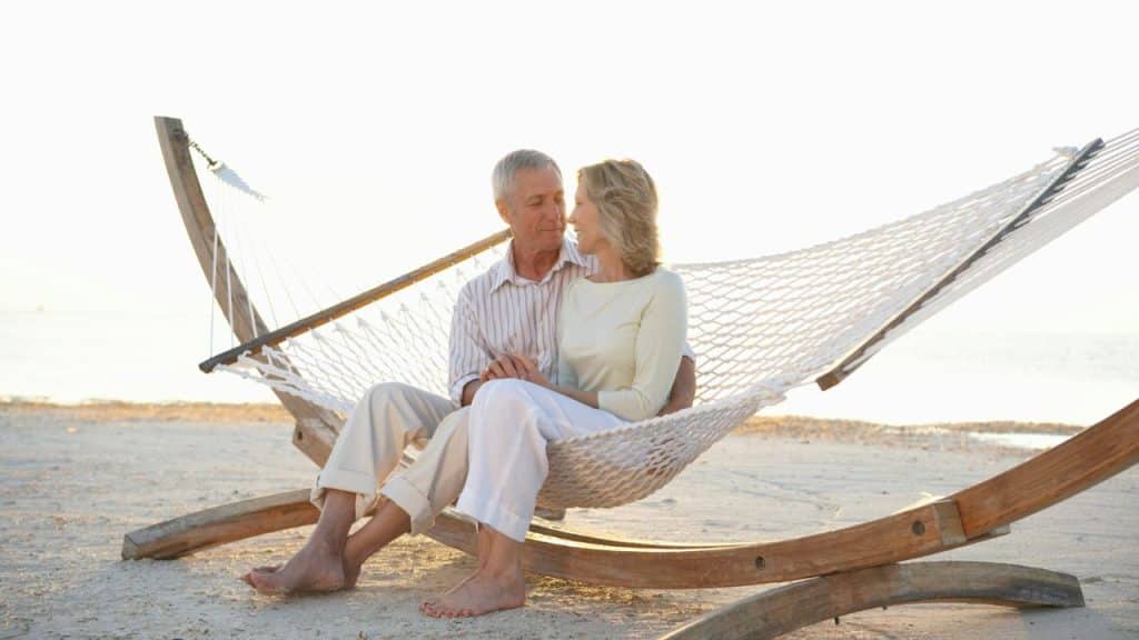 An older couple sitting closely together in a white hammock on a sandy beach.
