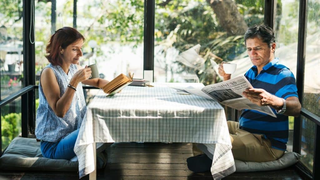 A man reading a newspaper and a woman reading a book at a glass-enclosed table.