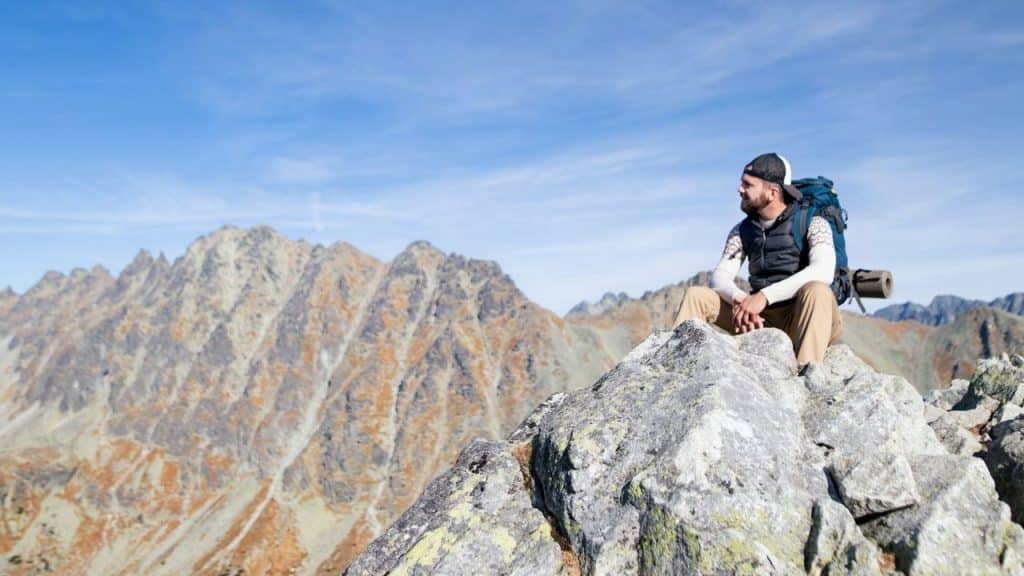 Hiker with a backpack sitting on a rocky mountain peak under a clear blue sky.