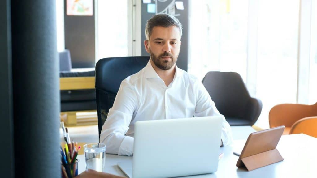 Man in a white dress shirt intently looking at a laptop on a white desk in an office.