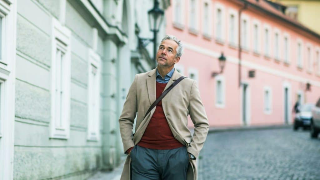Mature man in a tan coat and red sweater walking on a European city street with hands in pockets.